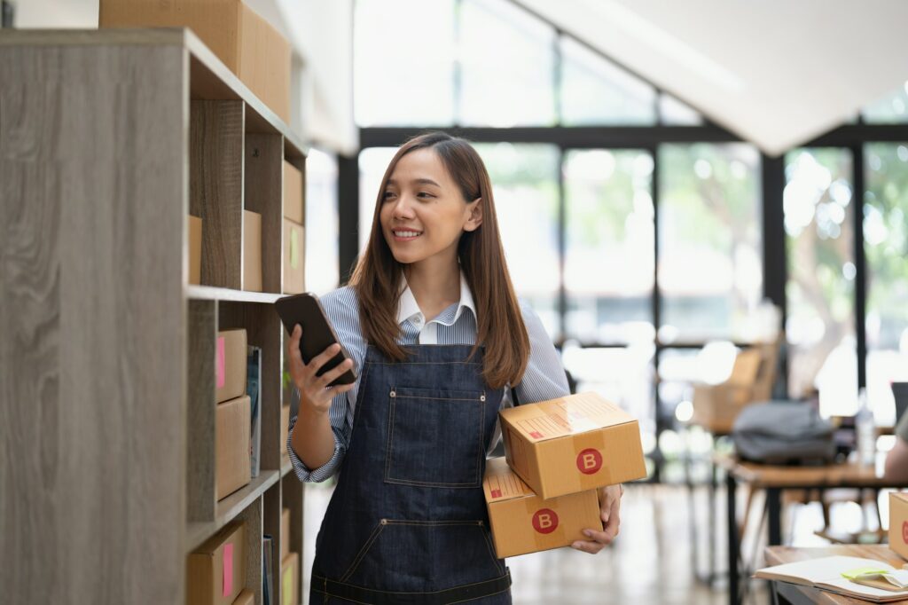 Attractive asian woman hold package box, using mobile phone checking stock order, startup SME