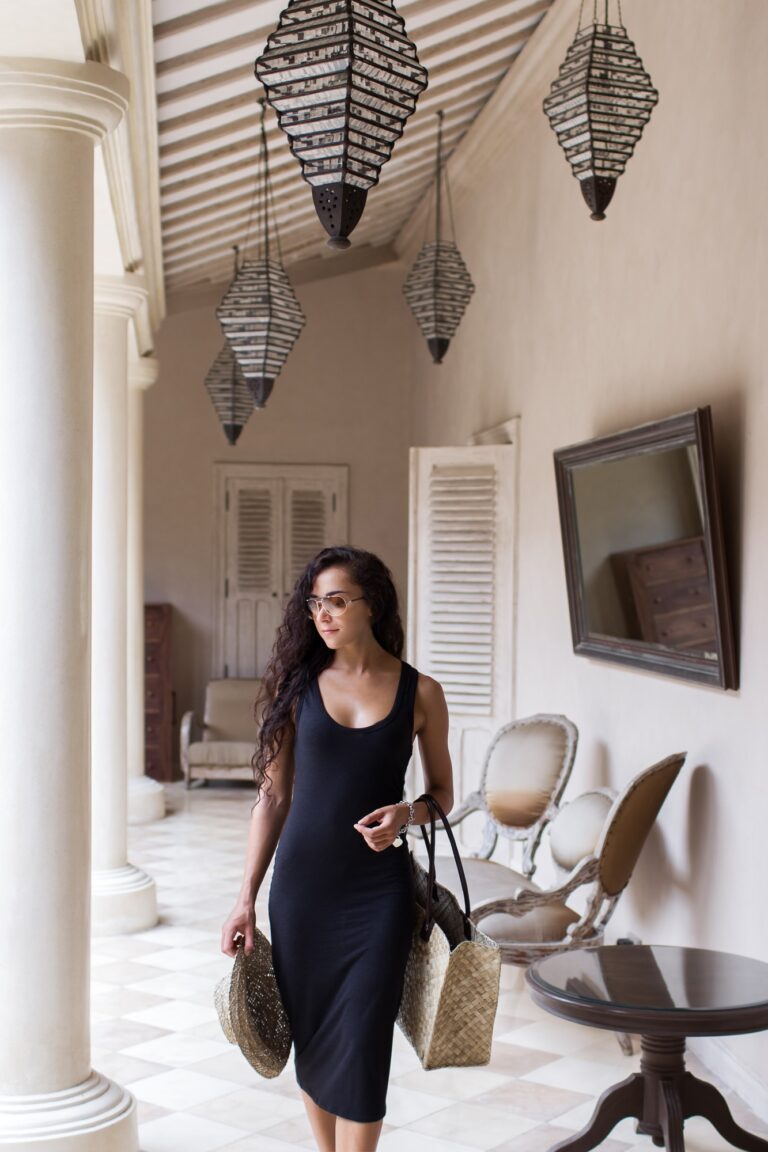 Pleased woman in black dress after shopping with straw bag in luxury mall with white walls