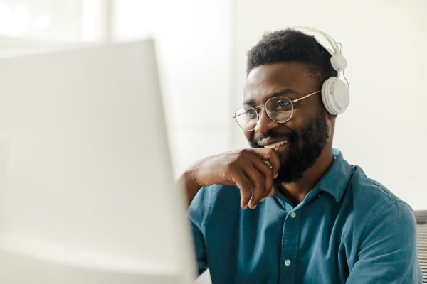 Happy black man in headphones working on computer, watching webinar, freelancing, taking part in